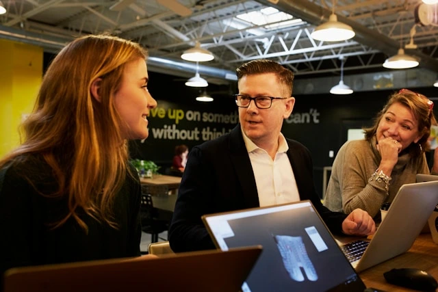 Three business professionals engaged in a focused discussion around laptops at a table, symbolizing collaborative strategy and client engagement in B2B sales or appointment generation.