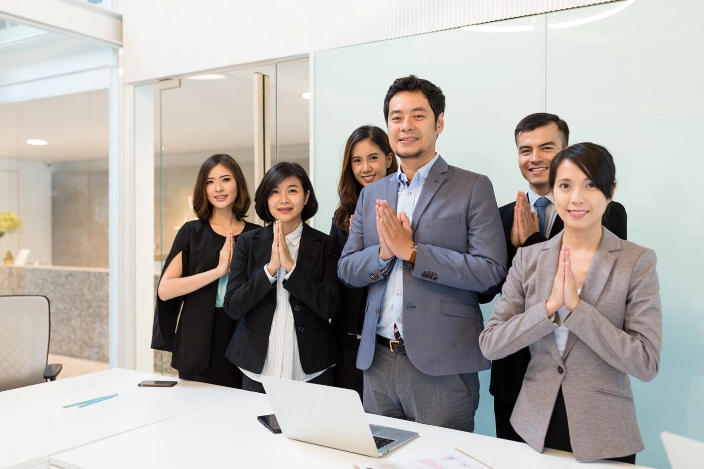 A group of smiling Thai business professionals in an office, performing the traditional 'Wai' greeting.
