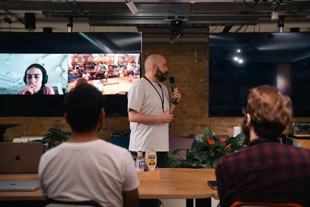 Hybrid business event with a speaker presenting in front of a small in-person audience, while virtual participants join on a large screen in the background.