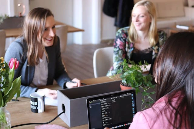Three professionals having a discussion around a desk with a laptop, plants, and coffee mugs, representing collaboration and partnership.